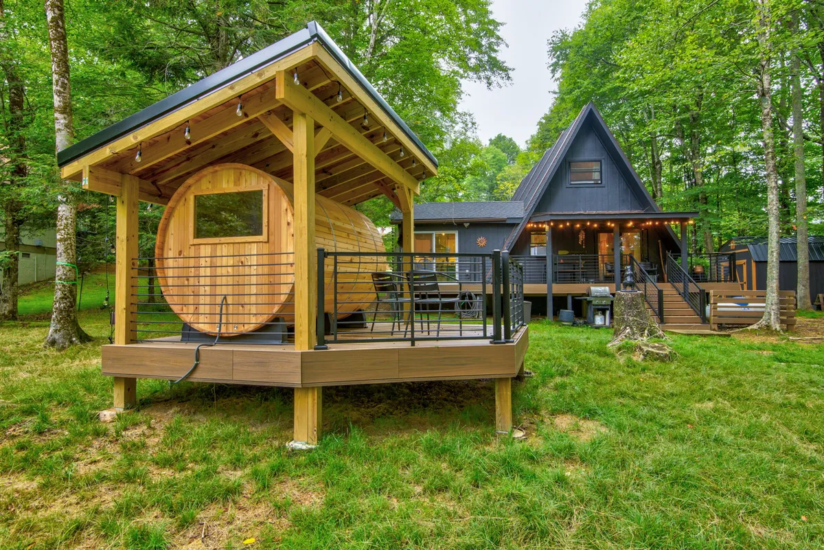 The hero image showcases a modern A-frame cabin surrounded by lush greenery, with a distinctive wooden sauna structure beside it. The scene evokes tranquility and retreat, featuring an inviting deck perfect for stargazing or sharing stories by the fire pit after a day exploring nearby nature trails.