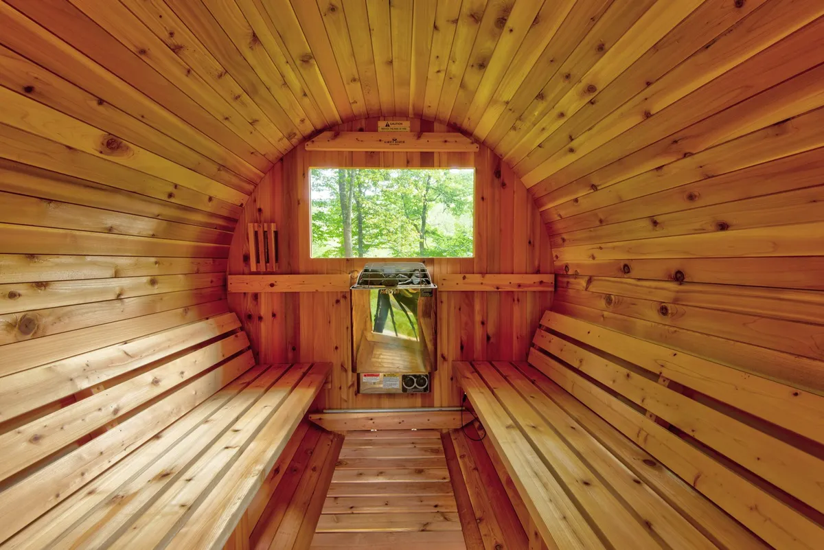 A calming interior shot of a wooden sauna with benches, a large window providing natural light, and a heating unit. The space is designed for relaxation and wellness after a day of mountain adventure.