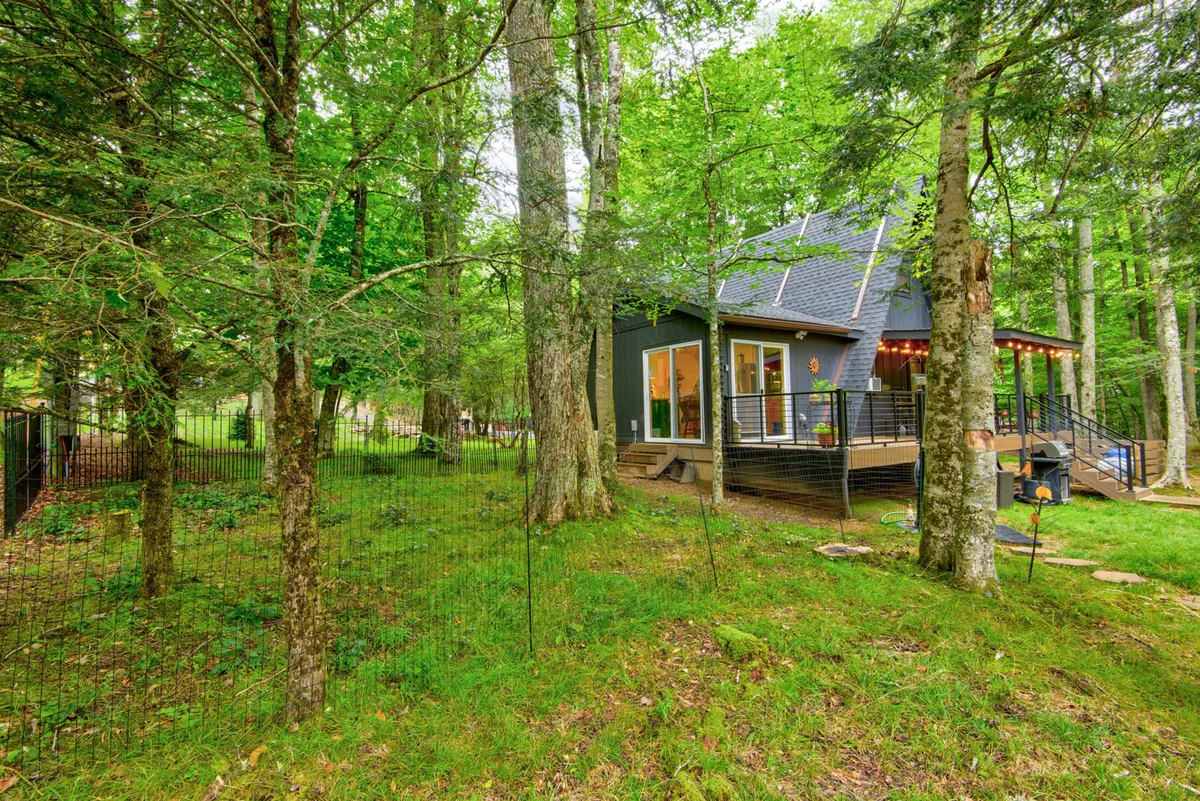 The photo highlights a modern cabin with a spacious deck and lush greenery all around. The deck area is set up for outdoor dining and relaxation, offering views of the surrounding landscape and a strong connection to nature.