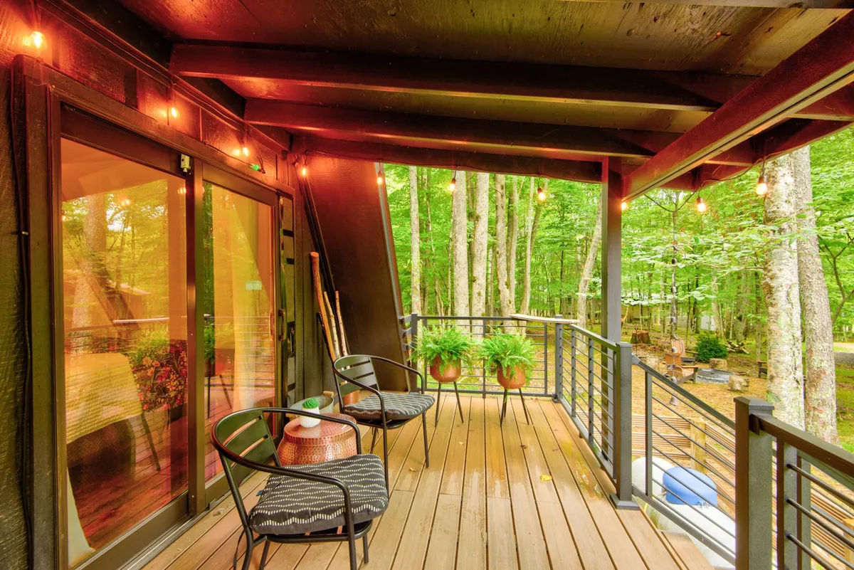 A peaceful exterior deck scene with hanging string lights, potted ferns, outdoor seating, and a backdrop of trees, ideal for morning coffee, evening relaxation, or social gatherings.