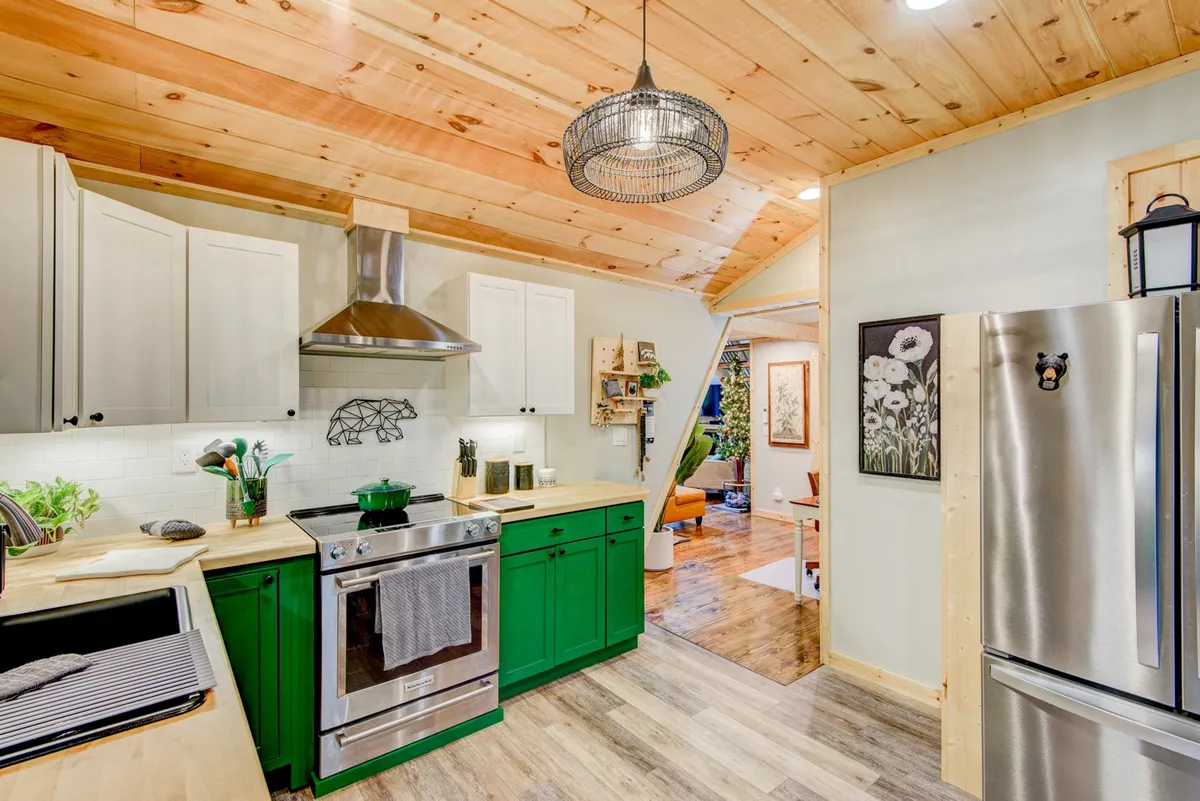 This image shows a cheerful kitchen featuring green cabinetry, stainless steel appliances, a wooden ceiling, and decorative wall art. The workspace is bright and inviting, with the stainless appliances clearly visible, and the overall scene reflects a fully equipped culinary space ideal for family meal preparation.