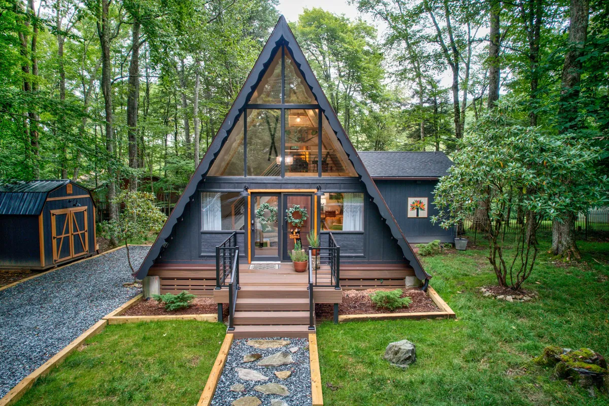 This exterior image features a striking A-frame structure with expansive glass windows, a spacious wooden deck, and a landscaped garden. The deck directly connects to the outdoor area, ideal for letting dogs enjoy the yard safely under supervision. The setting is tranquil, surrounded by nature, and hints at the pet-friendly, outdoor-focused lifestyle promoted in the section.