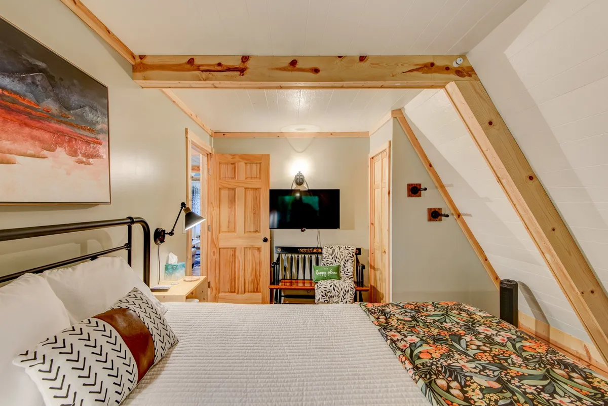 A warm, inviting bedroom with a comfortable bed adorned with patterned pillows, artwork on the wall, exposed wooden beams, a TV, and a seating area. The room is bathed in natural light, offering an ideal space for relaxation, watching TV, or enjoying a quiet morning coffee.