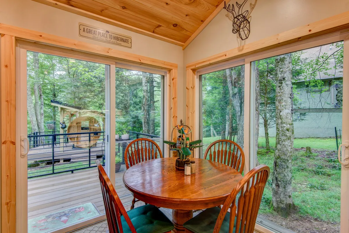 The image depicts a wooden dining table surrounded by large windows, with a decorative wall sign adding charm. The setting is ideal for family meals, morning coffee, or game nights, and the view suggests easy access to outdoor spaces for al fresco dining.
