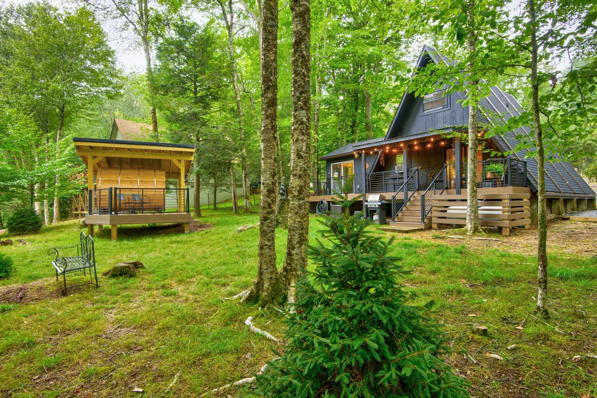 This image displays the A-frame house with a welcoming porch, a wooden gazebo perfect for gatherings, and a lush green lawn surrounded by trees. The setting is tranquil, with open grassy space that invites both children and pets to play safely, and plenty of room for outdoor family fun.
