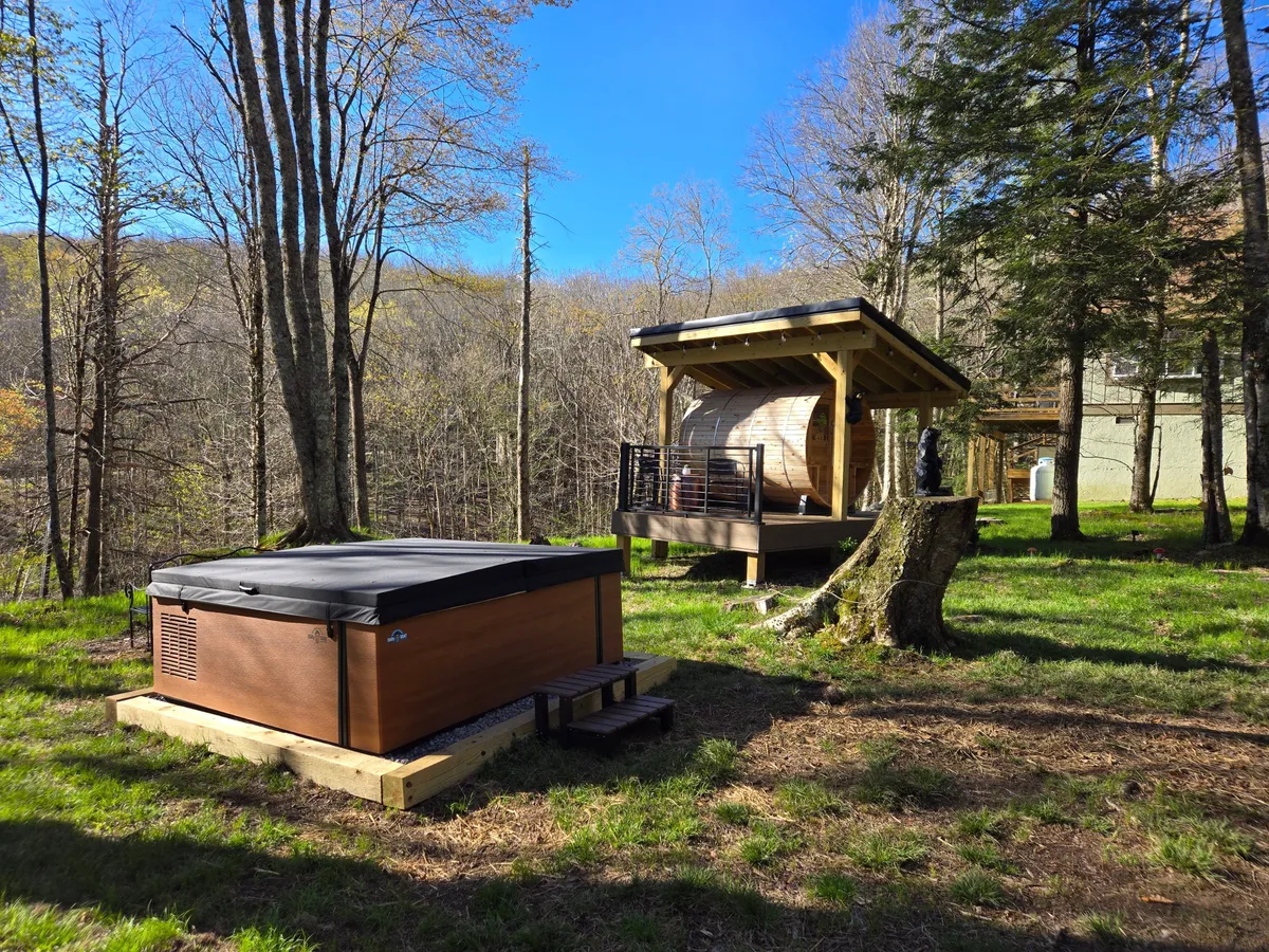 A serene exterior view featuring a traditional hot tub nestled on a wooden deck surrounded by lush trees. The setup is tranquil, with the hot tub taking center stage and hints of the deck's inviting space for relaxation after a day outdoors.