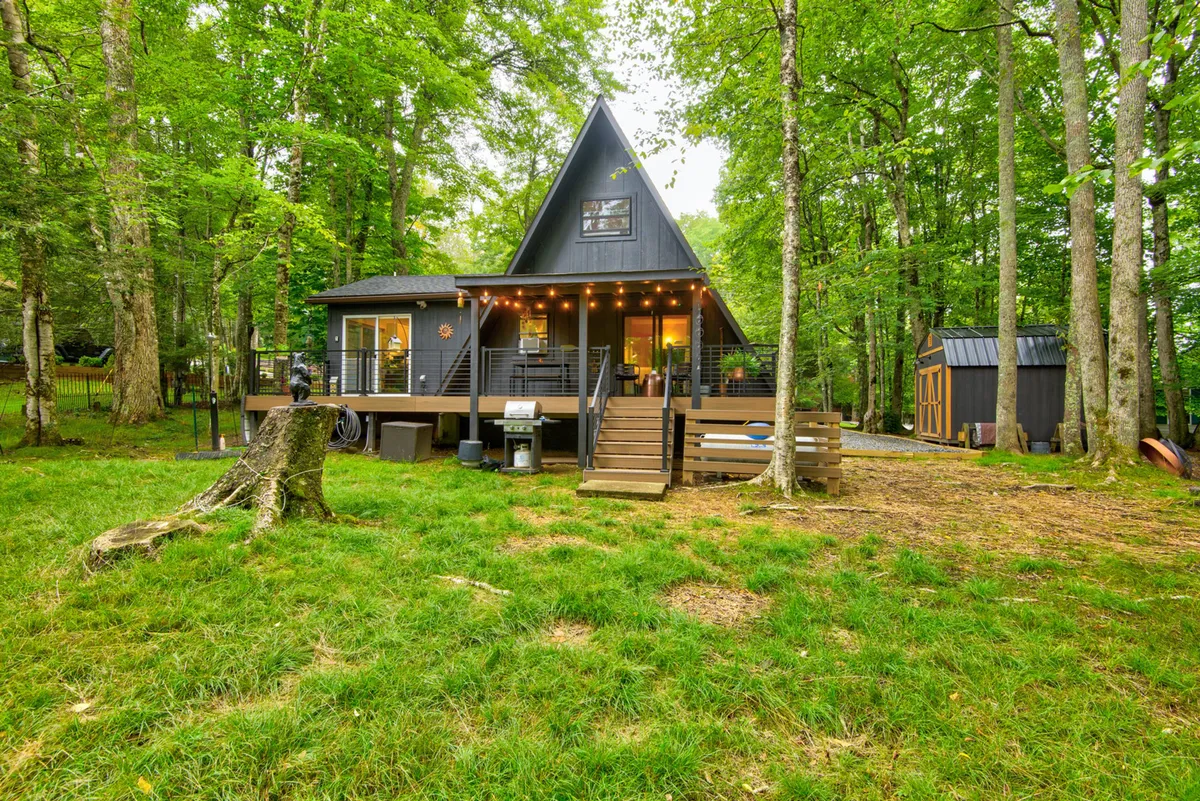 A cozy A-frame house stands amidst greenery, featuring a wooden deck with outdoor seating. The surrounding trees provide shade, while the deck serves as a comfortable spot for barbecuing, relaxing, or watching children play. The atmosphere is serene, making it ideal for families returning from outdoor activities.