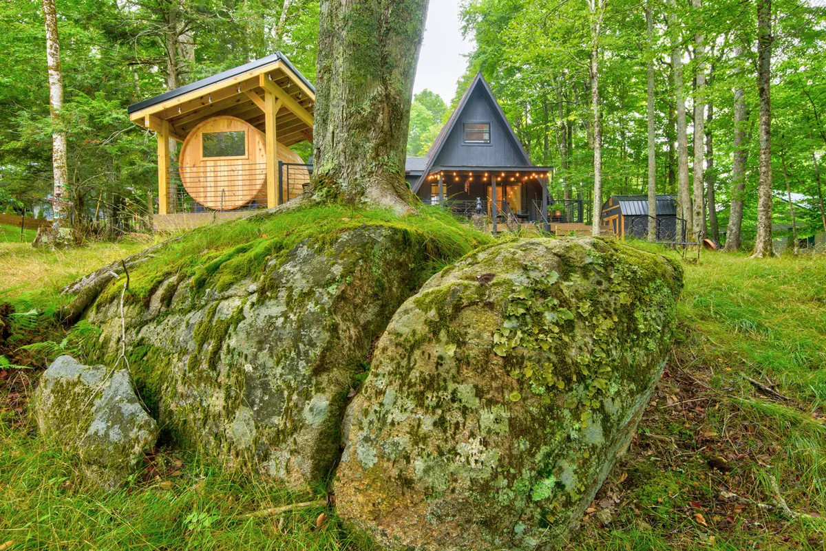 This image shows a charming wooden cabin with a distinctive circular window and an A-frame house, nestled among tall trees and a large moss-covered rock. The scene exudes tranquility and offers a glimpse of the natural surroundings, inviting guests to explore the woods or enjoy outdoor gatherings.