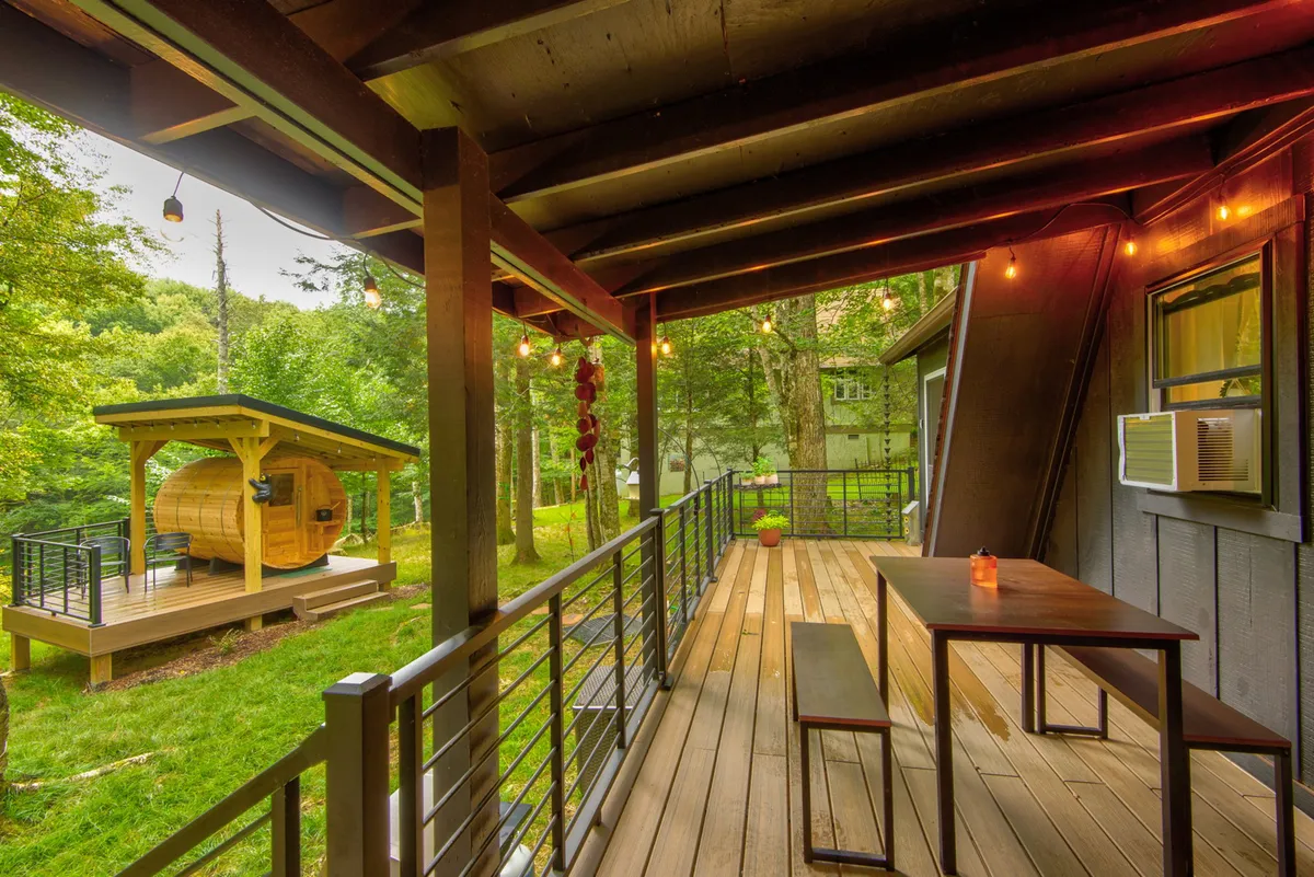 A cozy deck scene with a barrel sauna prominently displayed beside a dining table under string lights. The surrounding trees and soft lighting evoke a sense of relaxation, social connection, and wellness.