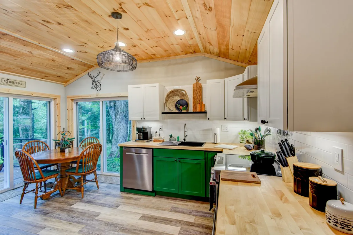 The image highlights the kitchen and dining area, with green kitchen cabinets, a wooden dining table set for four, and large windows overlooking lush greenery outside. The scene exudes warmth and natural light, emphasizing the ease of gathering for meals and the cozy, inviting feel of the dining area.