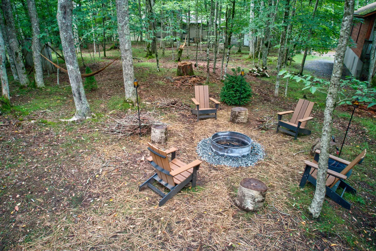 A cozy exterior setting featuring a fire pit encircled by wooden chairs and a hammock, all set amidst tall trees. This communal space invites guests to gather, share stories, and stargaze in the crisp mountain air.
