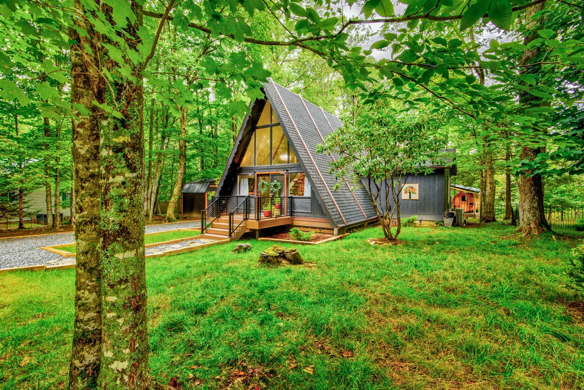An inviting view of the A-frame cabin’s exterior with a welcoming front porch and steps, surrounded by lush greenery. The scene is tranquil and rejuvenating, linking the property’s architecture to its natural mountain environment.